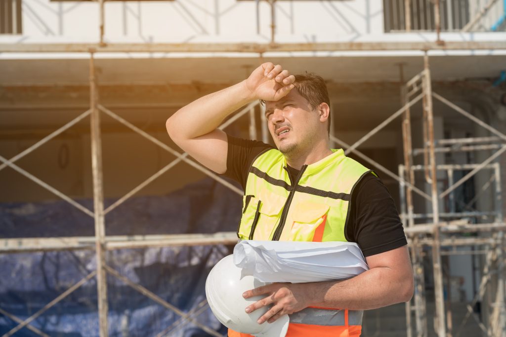 Construction worker in a high-visibility vest on a scaffolded site, holding a white hard hat.